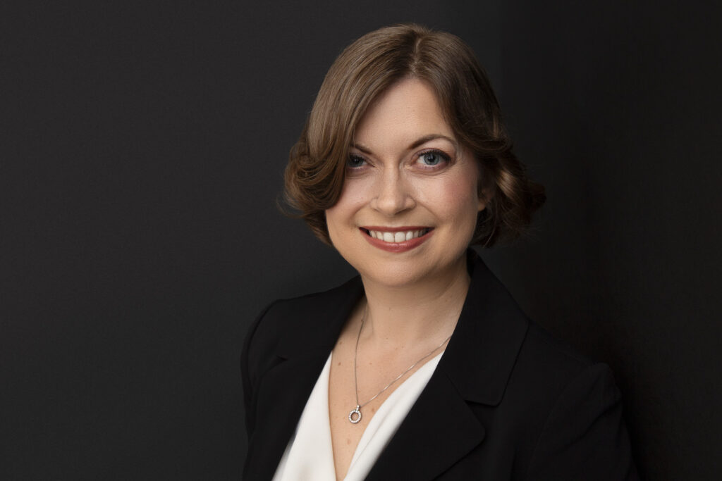 Personal branding headshot of smiling woman with short black hair in a white top with black jacket on a black background.