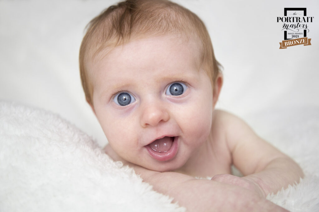 Infant holding its head up and smiling at the camera. Infant is propped up on a fuzzy white blanket.